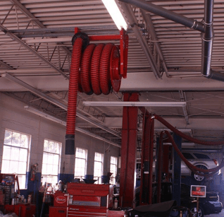 Exhaust hose reel shown installed in a repair shop with overhead exhaust removal systems and vehicles in the background.